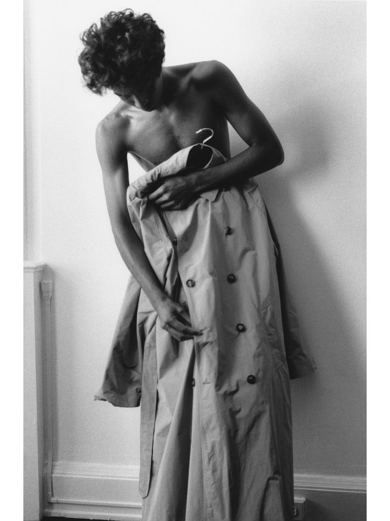 Shirtless man with curly hair putting on a double-breasted trench coat indoors