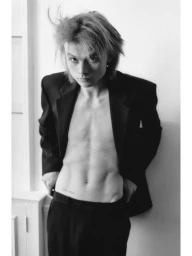 Monochrome portrait of a young man wearing an unbuttoned black blazer against a plain white interior wall