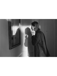 Monochrome image of a person standing in profile looking at a wall mirror with a strong shadow cast behind them.