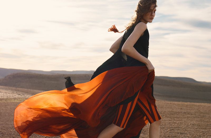 Model in flowing black dress and striped orange skirt walking across open terrain under an overcast sky.