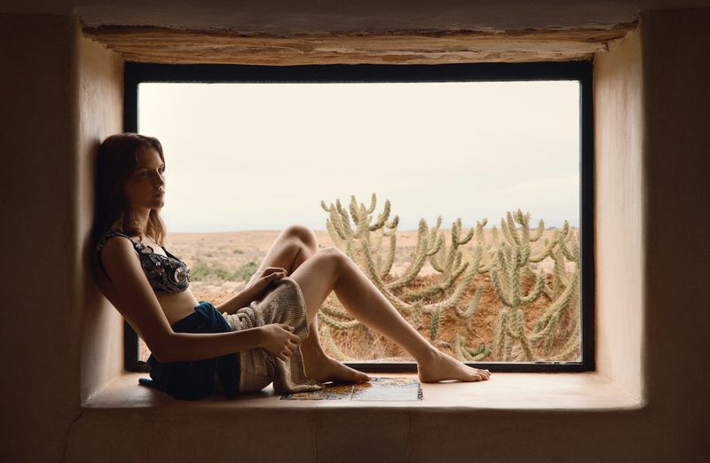 Model seated inside a wide window frame overlooking an arid desert landscape with cacti for Vogue Thailand.