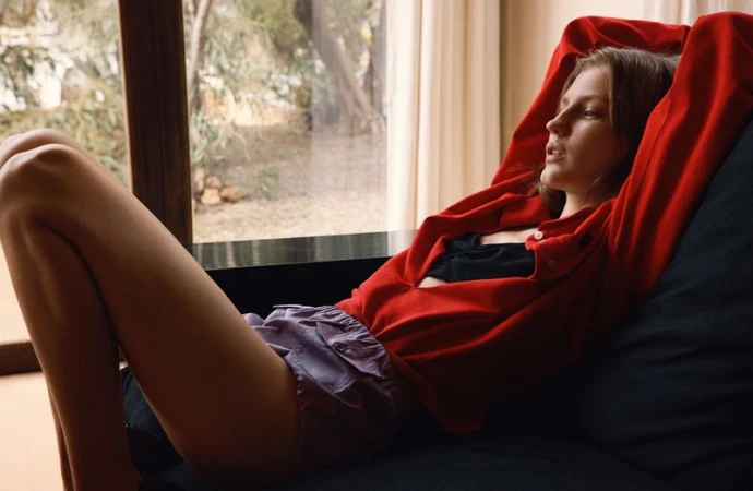 Model relaxing on a dark sofa wearing a red shirt and purple shorts with arms raised against a bright window.