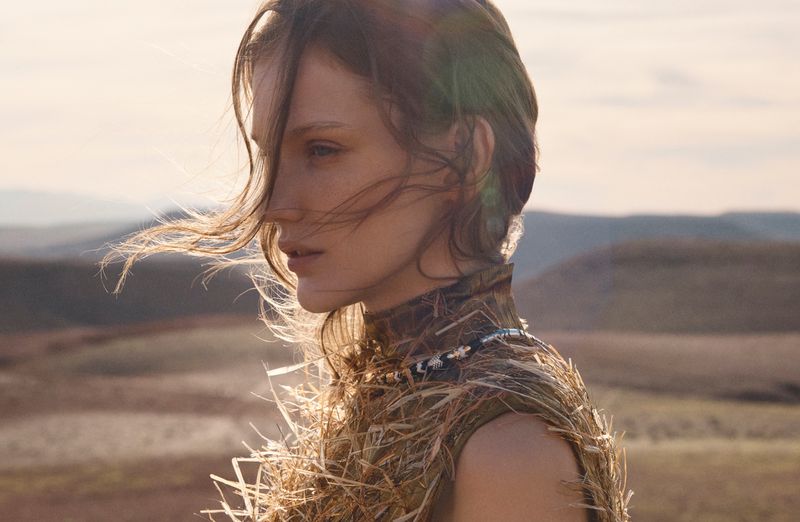 Profile portrait of a female model with windswept hair wearing a textured straw-like garment against an open landscape.