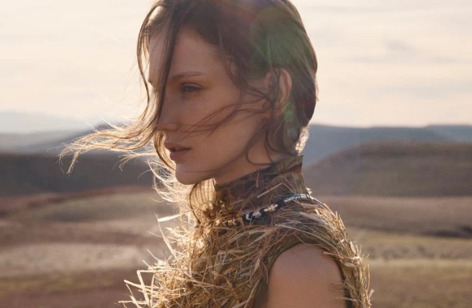 Profile portrait of a female model with windswept hair wearing a textured straw-like garment against an open landscape.