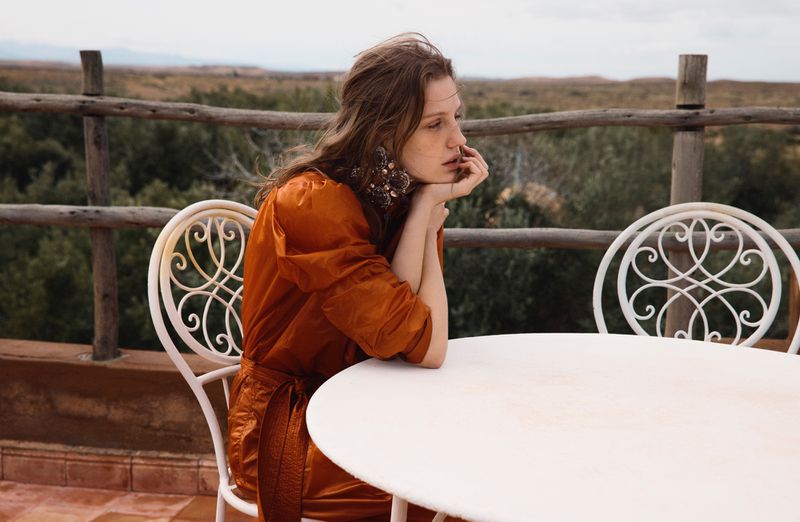 Model in an orange dress seated at a white table outdoors against a rustic wooden fence and distant landscape