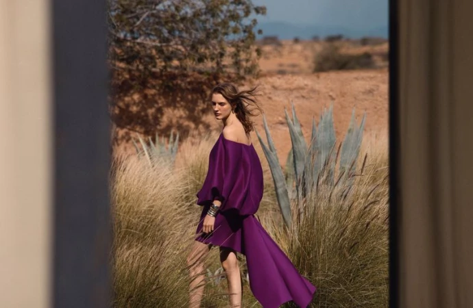 Model wearing an off-the-shoulder purple dress standing amidst tall grass in an arid landscape with distant hills