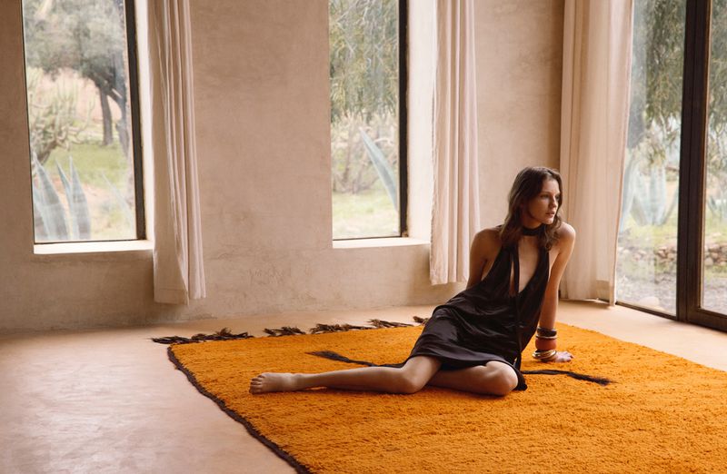 Model wearing a black halter dress seated on a bright orange rug inside a sunlit room with large windows.