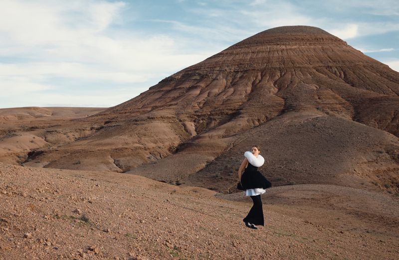 Model standing in a vast desert landscape wearing a black outfit with a large white feathered collar