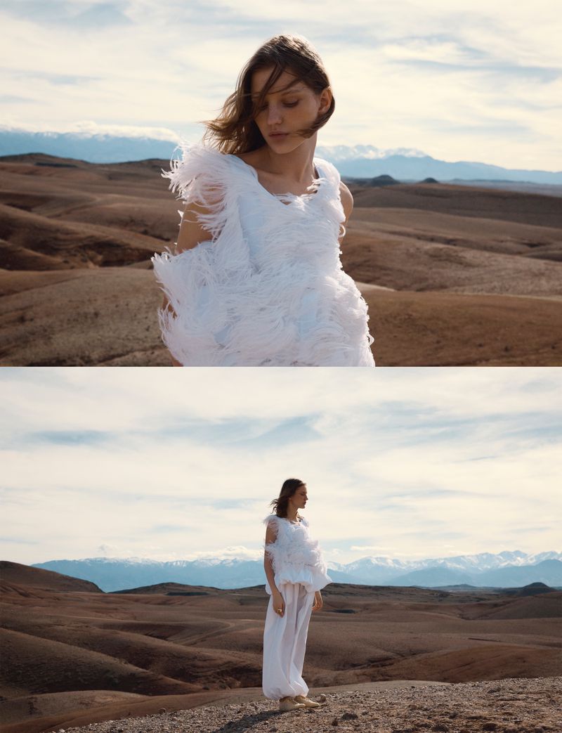 Model in a white feathered top and trousers standing in a barren desert landscape with distant mountains under a cloudy sky.