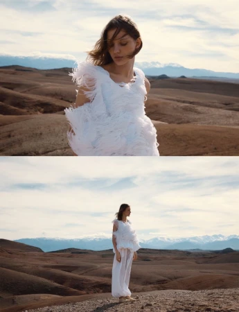 Model in a white feathered top and trousers standing in a barren desert landscape with distant mountains under a cloudy sky.