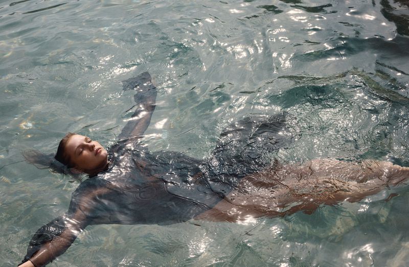 Model floating flat on her back in crystal-clear water with bright sunlight shimmering across the surface.