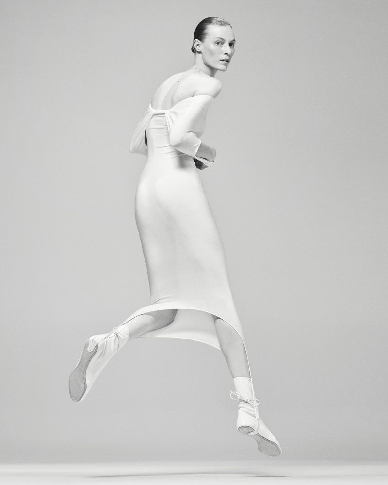 Model Julia Nobis leaping in a white off-the-shoulder dress against a seamless light grey studio background.