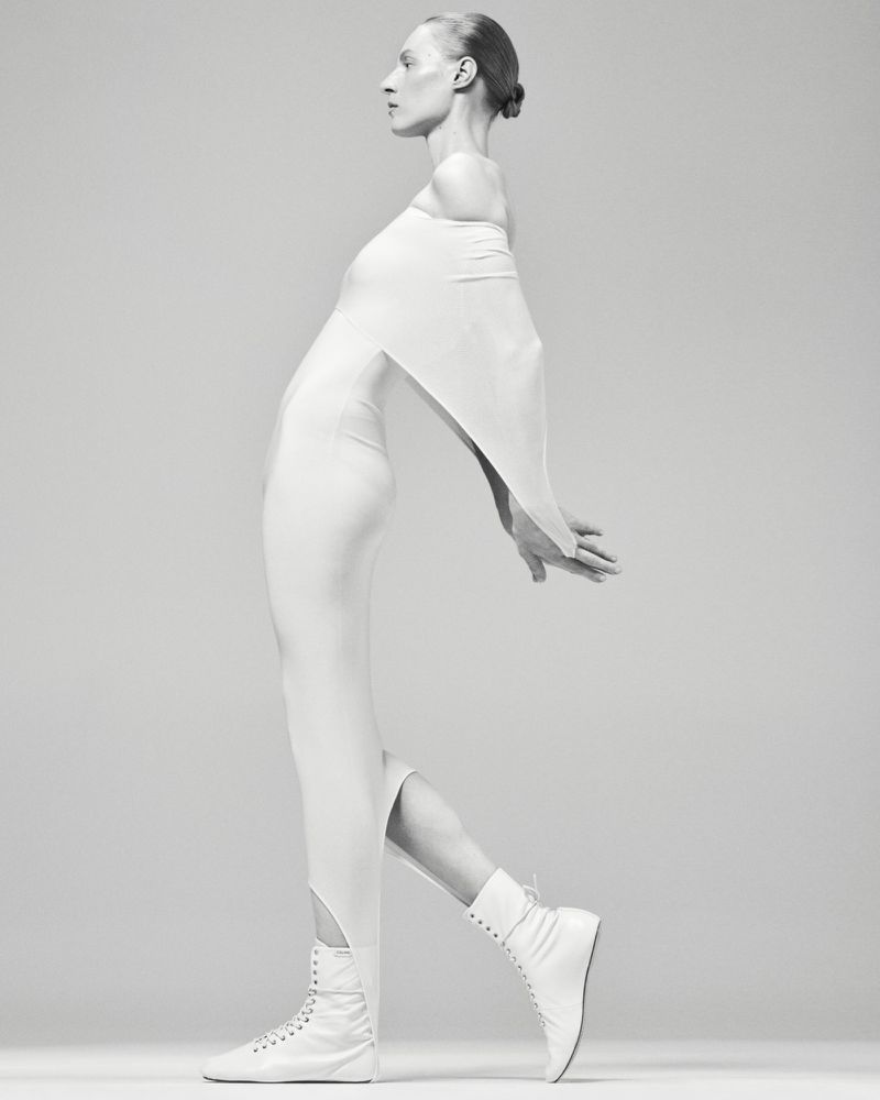 Model Julia Nobis wearing a white off-shoulder dress and white boots against a grey seamless background.