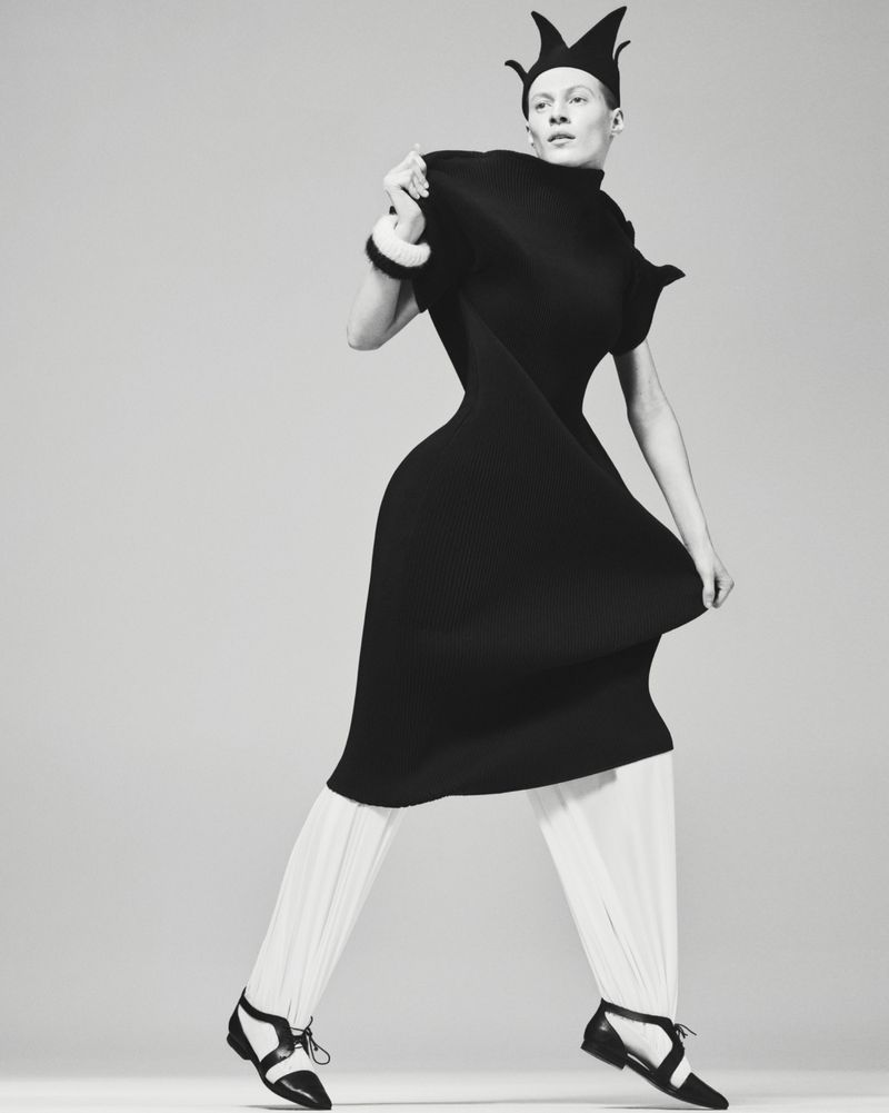 Fashion photo of a model in a black dress and crown-like hat with white pants and black shoes, striking a pose against a neutral backdrop.