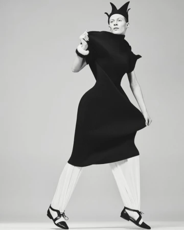 Fashion photo of a model in a black dress and crown-like hat with white pants and black shoes, striking a pose against a neutral backdrop.