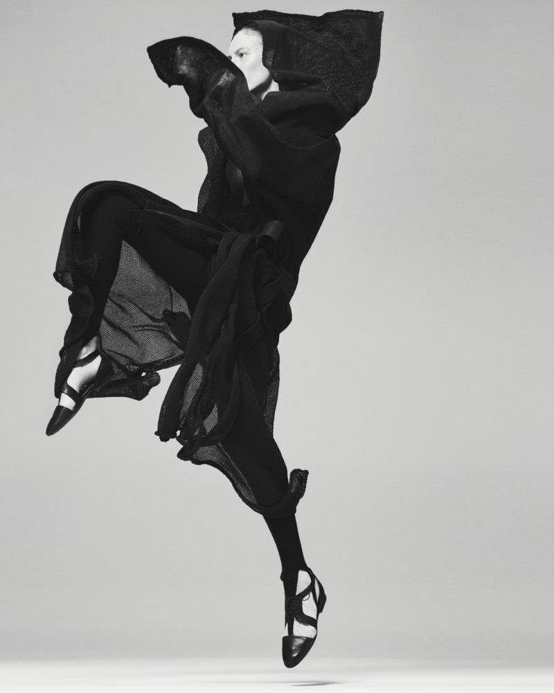 Model Julia Nobis leaping mid-air in a hooded black dress and strapped shoes against a plain grey backdrop.