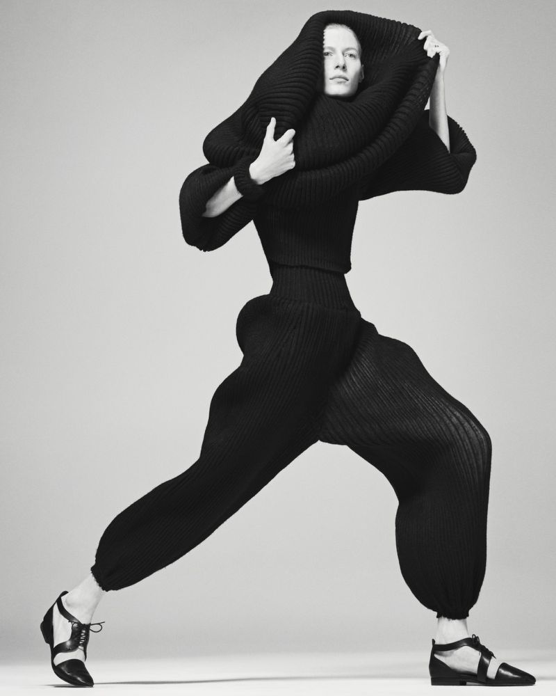 Model Julia Nobis in a sculptural black knit outfit and dark shoes against a light grey background