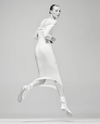 Model Julia Nobis leaping in a white off-the-shoulder dress against a seamless light grey studio background.