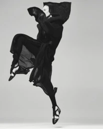 Model Julia Nobis leaping mid-air in a hooded black dress and strapped shoes against a plain grey backdrop.