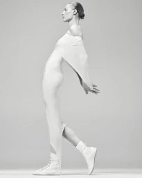 Model Julia Nobis wearing a white off-shoulder dress and white boots against a grey seamless background.