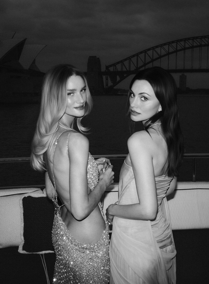 Two women in evening dresses posing outdoors at night with the Sydney Harbour Bridge visible in the background.