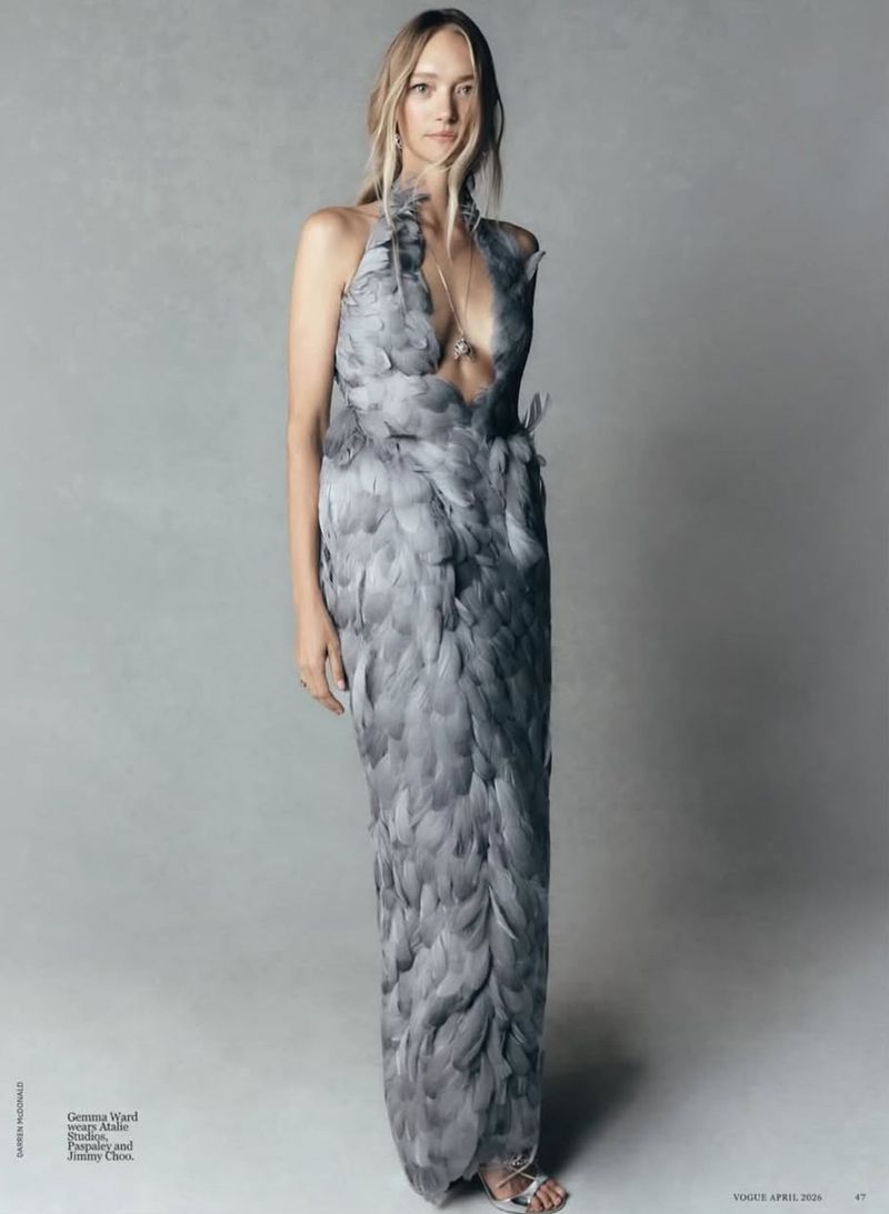Fashion editorial shot: A slender woman poses gracefully in a floor-length gown made of grey feathers, set against a neutral grey backdrop.