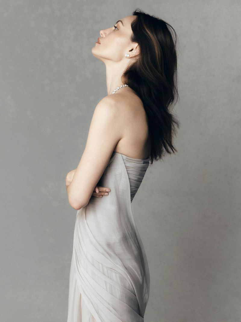 Profile portrait of a woman wearing a strapless grey dress and pearl jewelry against a neutral grey studio background