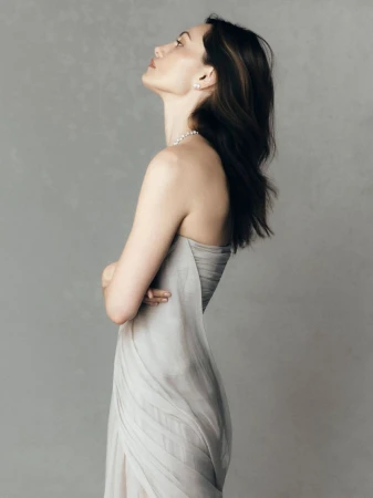 Profile portrait of a woman wearing a strapless grey dress and pearl jewelry against a neutral grey studio background