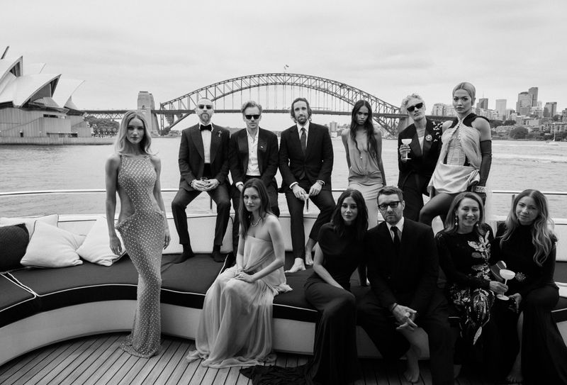 Group portrait of famous actors and models in formalwear on a yacht with the Sydney Harbour Bridge and Opera House in the background.