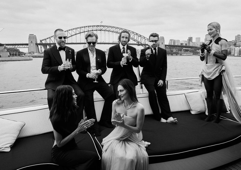 Group of formal-wearing figures on a boat deck with the Sydney Harbour Bridge visible in the background