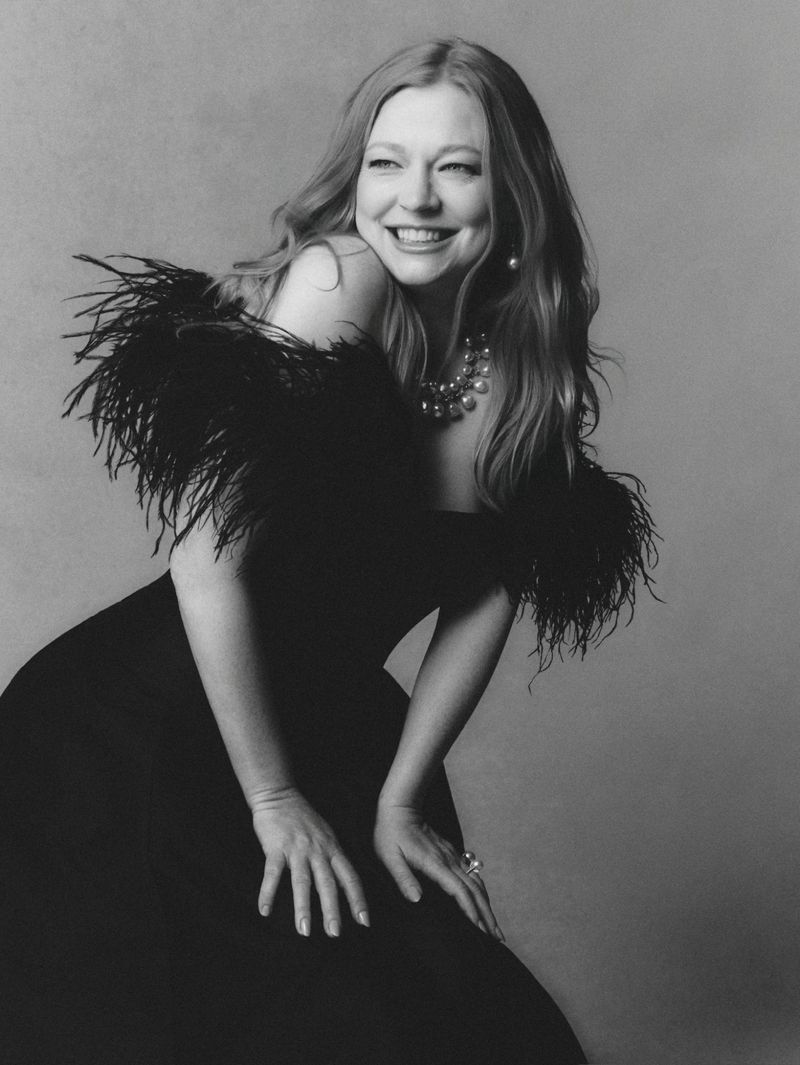 Portrait of Sarah Snook wearing a black dress with feather trimmed shoulders, smiling against a grey background