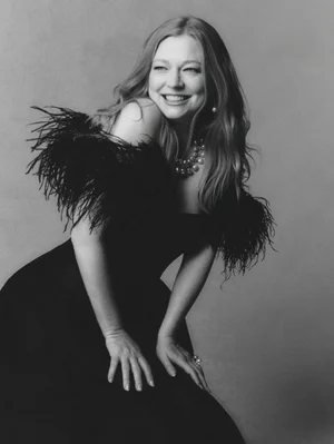 Portrait of Sarah Snook wearing a black dress with feather trimmed shoulders, smiling against a grey background