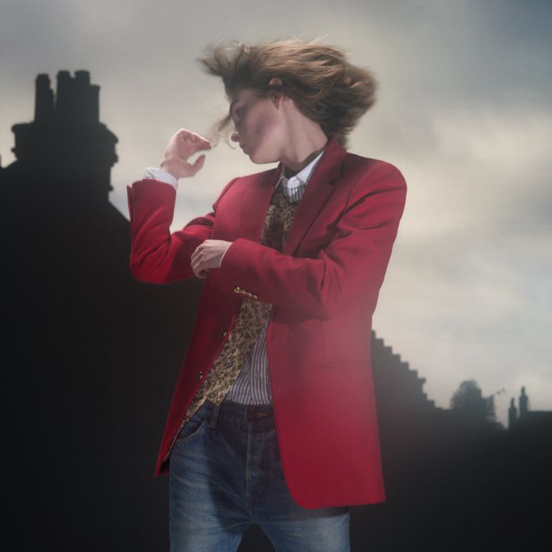 Model with blurred hair wearing a red blazer and jeans against a moody outdoor building silhouette background for Pop Magazine #54.