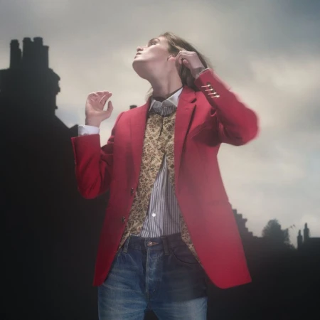 Model in red blazer and patterned vest looking upward against an overcast sky and chimney silhouettes