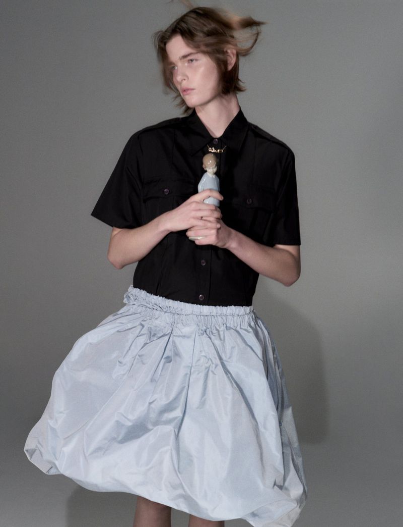 Model with medium-length brown hair wearing a black short-sleeve shirt and pale blue skirt against a neutral grey seamless background