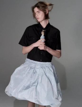 Model with medium-length brown hair wearing a black short-sleeve shirt and pale blue skirt against a neutral grey seamless background