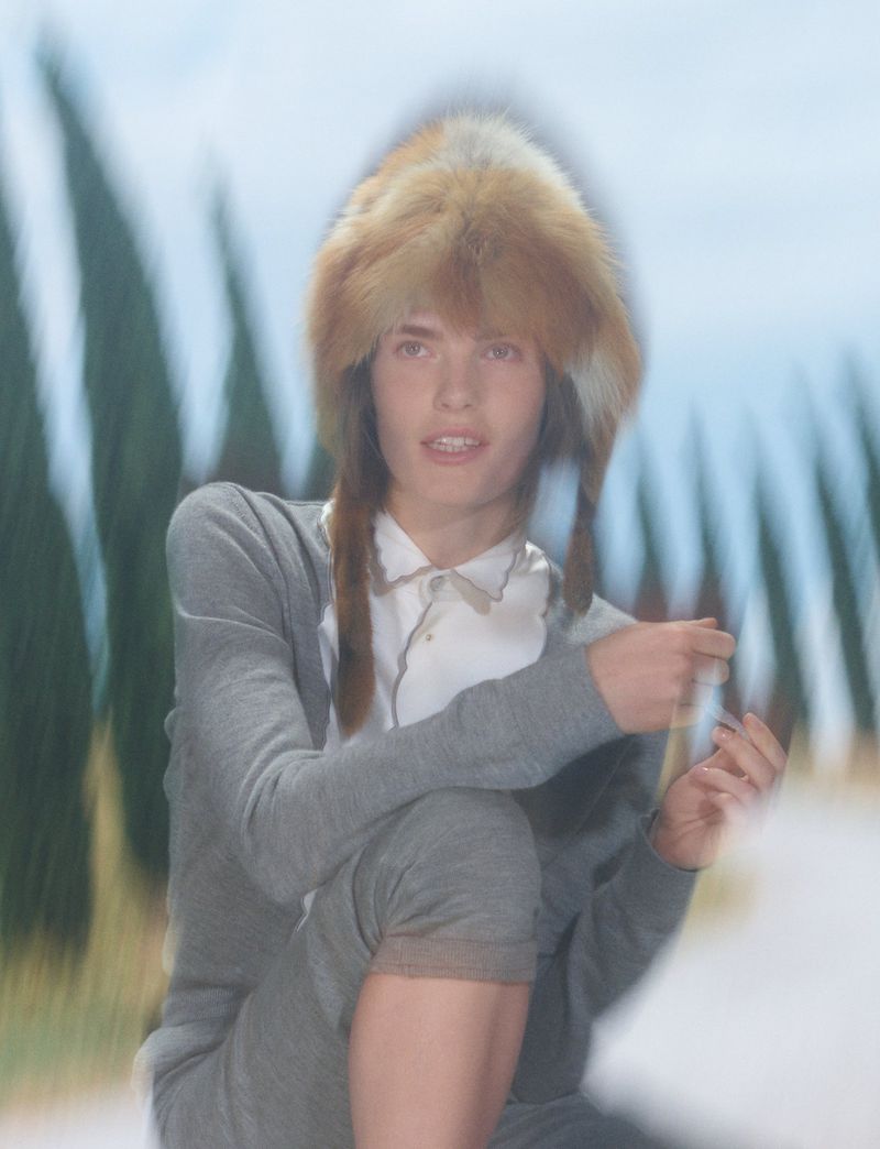 Model wearing a fuzzy fur hat and grey knit sweater posing outdoors with blurred trees in the background for Pop Magazine