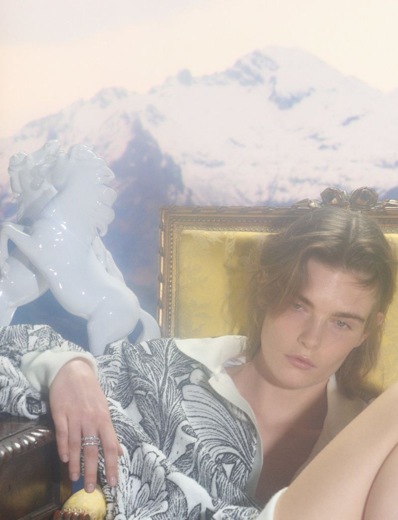 Model seated on an ornate chair wearing a patterned white top against a mountain backdrop with a white horse sculpture