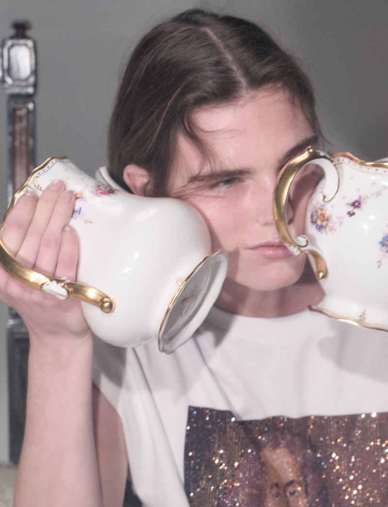 Model holding two porcelain teapots to ears wearing white graphic t-shirt against neutral grey background
