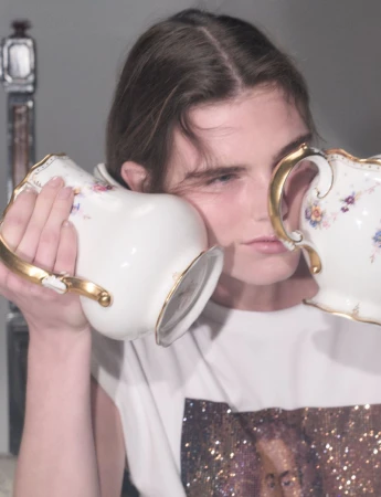 Model holding two porcelain teapots to ears wearing white graphic t-shirt against neutral grey background
