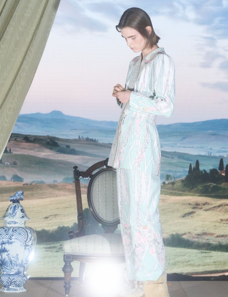 Model standing in a patterned pale blue two-piece outfit beside an antique chair against a scenic painted landscape backdrop.