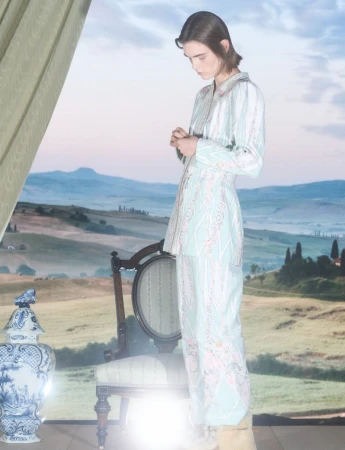 Model standing in a patterned pale blue two-piece outfit beside an antique chair against a scenic painted landscape backdrop.