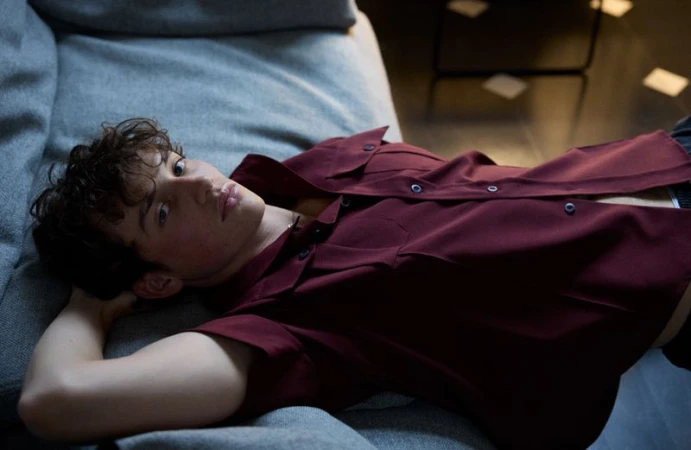 A young male model lying on a grey fabric sofa wearing an unbuttoned burgundy shirt for a 10 Men Magazine editorial.