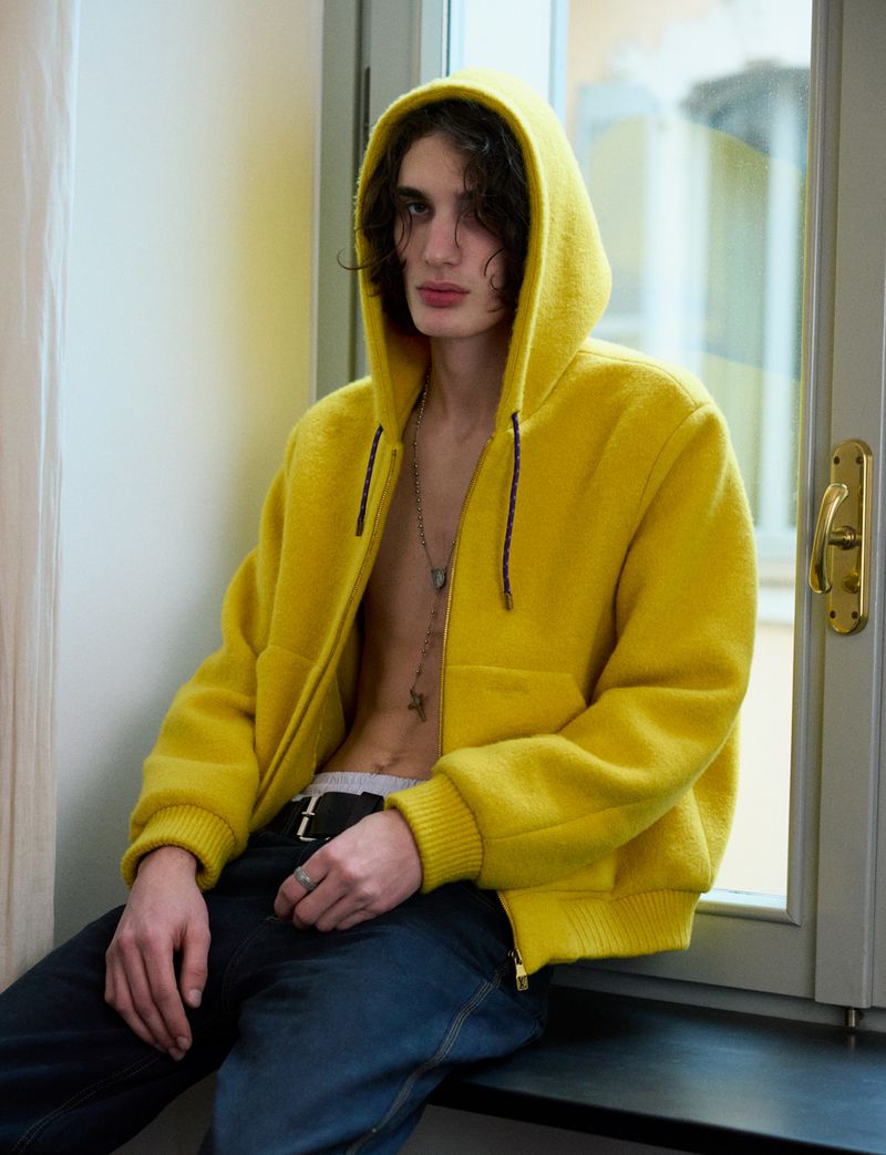 A young male model wearing a bright yellow hooded jacket and dark trousers seated by a window.
