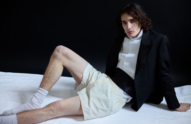 Model seated on a white surface wearing a black blazer, white shirt, cream shorts, and white socks against a dark background