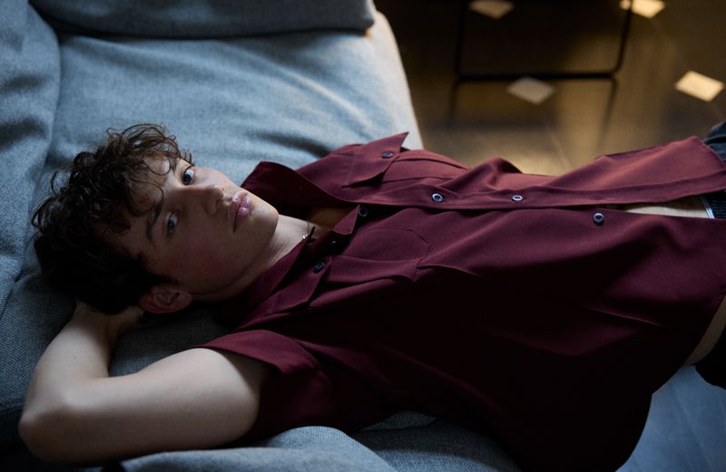 A young male model lying on a grey fabric sofa wearing an unbuttoned burgundy shirt for a 10 Men Magazine editorial.