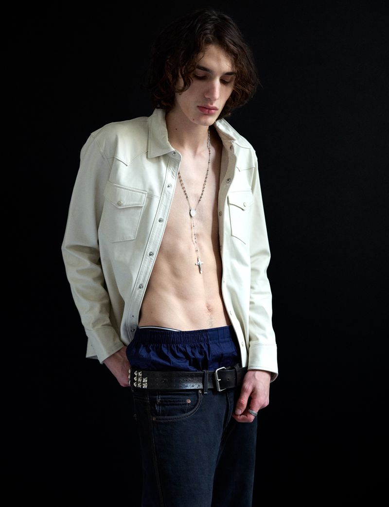 A young male model wearing an unbuttoned cream shirt, dark jeans, and a silver necklace against a black background.