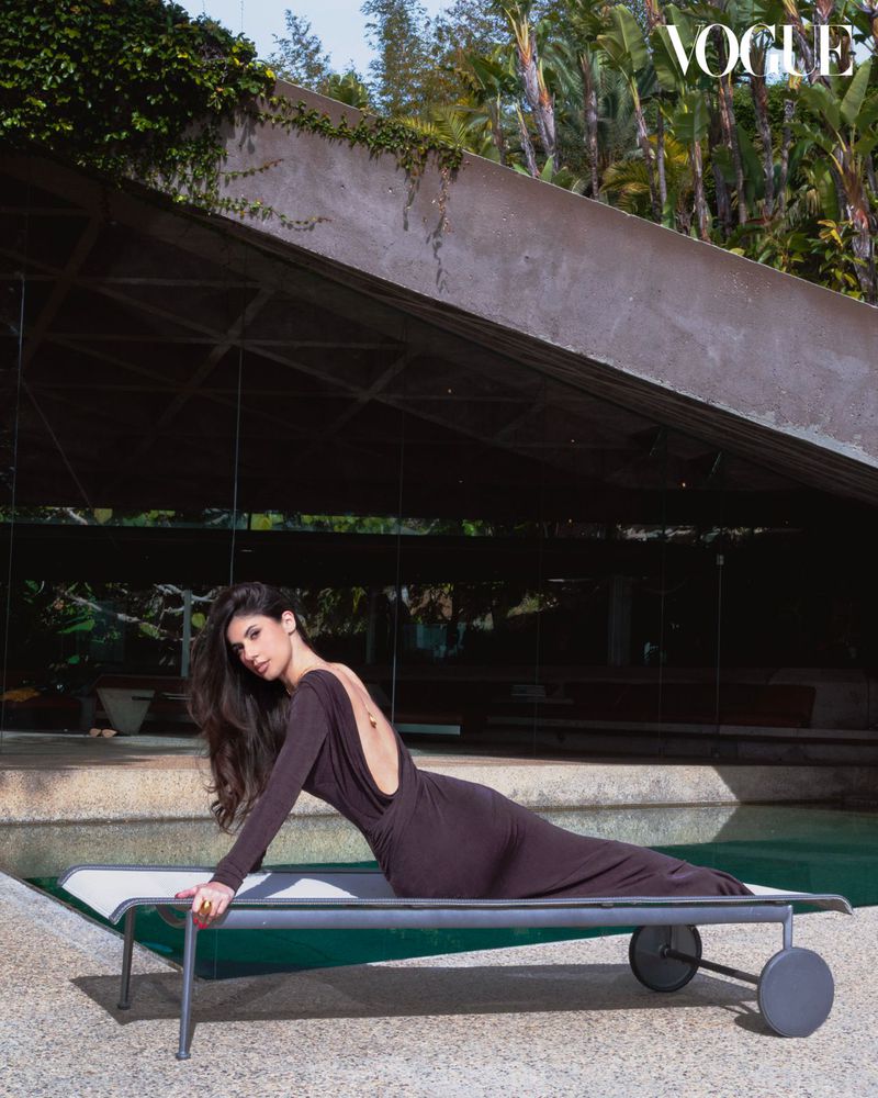 Woman in long-sleeve dress lounges on chaise lounge by pool in modern architectural setting with tropical foliage, Vogue logo visible.