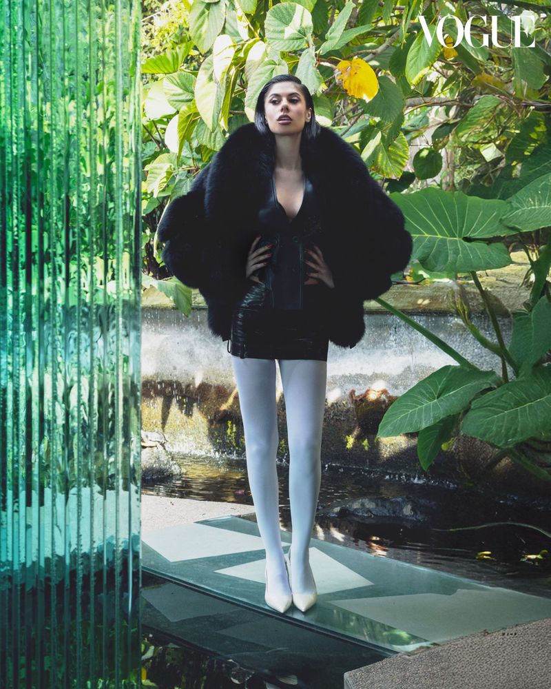 Model Ines Trocchia wearing a black faux fur coat and white tights standing outdoors among lush green tropical plants.