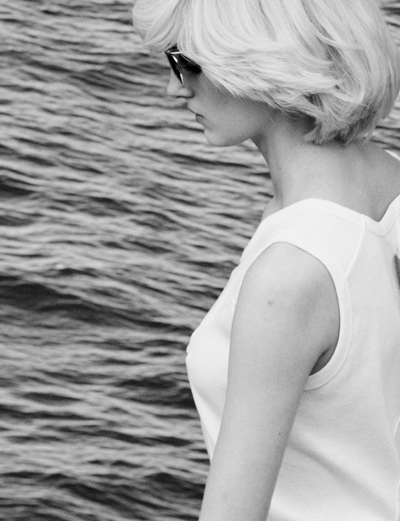 Monochrome portrait of a blonde model wearing a white top and sunglasses looking over rippled water for Pop Magazine